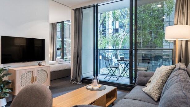 A modern living room with a flat-screen TV, a wooden coffee table, a grey sofa, and a large window offering a view of the outdoors. - StayCentral - Oasis on Mackenzie (Melbourne)