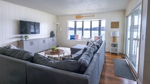 A modern living room with a grey sofa, a flat-screen TV, a wooden paddle wall decor, a large window with a view of a lake, and a small side table with a lamp.