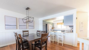 A dining area with a wooden table and chairs, a kitchen in the background, and artwork on the walls.