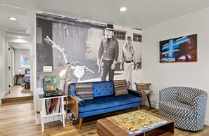 A living room with a blue sofa, a patterned armchair, a coffee table with a puzzle, a bookshelf with books and a vintage radio, and a large black and white mural on the wall. - Dapper Duo - 2 Adjacent Homes w/Fully Fenced Yards, AC, WD, Fast WiFi, Smart TVs (Flagstaff)