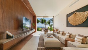 Een moderne woonkamer met een grote flatscreen televisie, een zithoek, een salontafel en uitzicht op palmbomen en de oceaan door grote ramen.