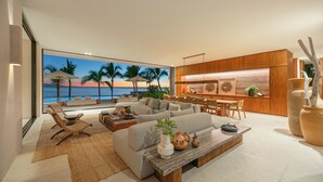 Een moderne woonkamer met een groot raam dat uitkijkt op een strand, een gezellige zithoek en een keuken met houten elementen.