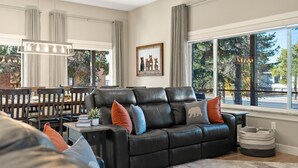 Un salon moderne avec un canapé en cuir, une table à manger et des chaises. Une grande fenêtre offre une vue sur les arbres et la rue.