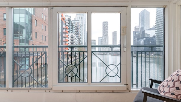 Een kamer met een groot raam dat uitkijkt op een stadsgezicht, een houten vloer en een zwarte bank met een gestreept kussen.