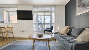 Ein modernes Wohnzimmer mit einem grauen Sofa, einem runden Couchtisch, einem Korbstuhl und einem an der Wand montierten Fernseher.