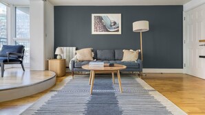 Ein modernes Wohnzimmer mit dunkelblauer Akzentwand, einem runden Couchtisch, einem grauen Sofa mit Kissen und einem schwarzen Sessel, sowie einer Stehlampe.