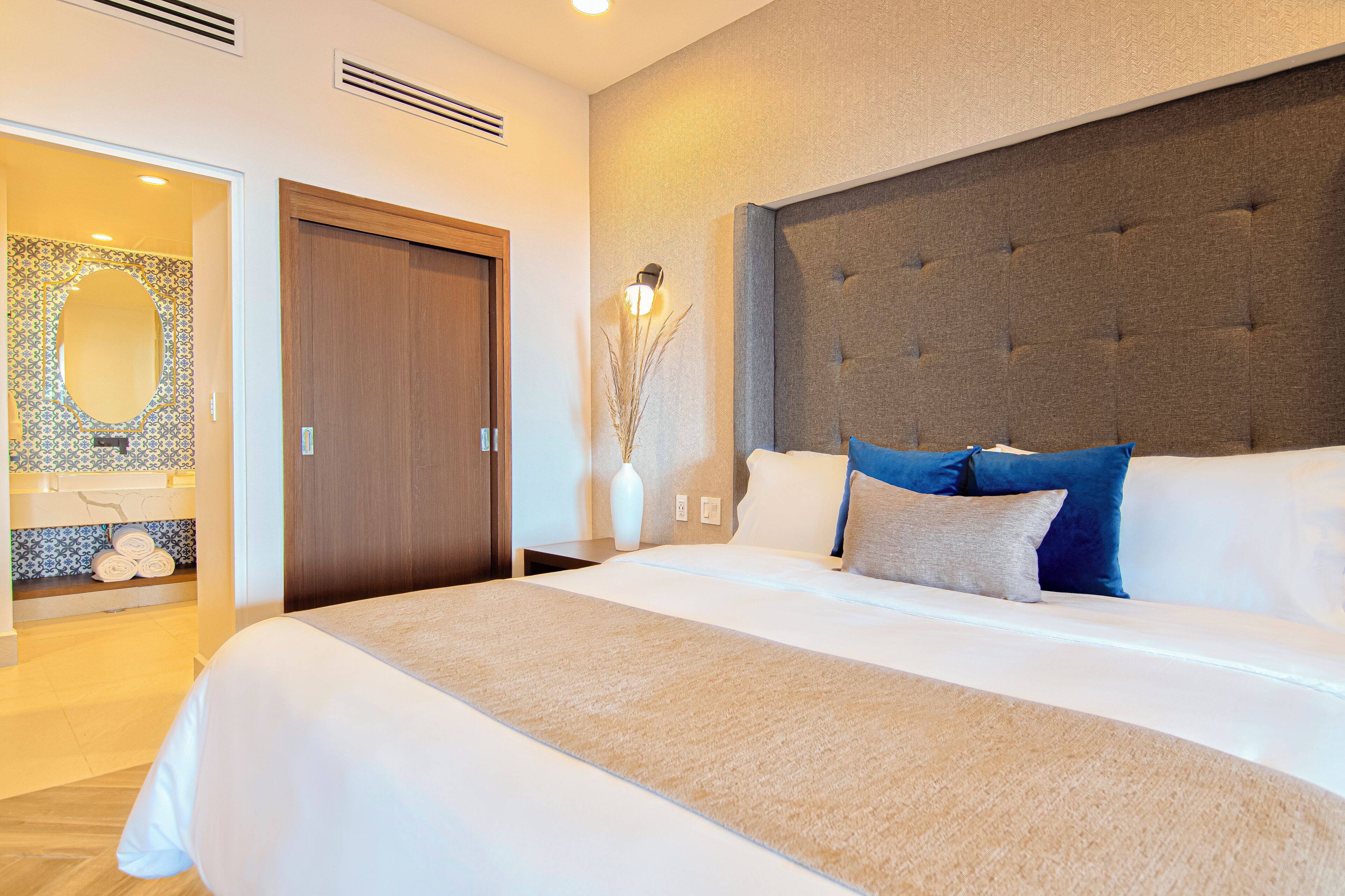 Una habitación de hotel moderna con una cama grande, una puerta de madera, un baño con espejo y salpicadero de azulejos, y un aplique de pared.