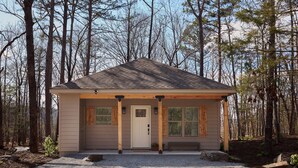 A small, single-story cabin with a front porch, a door, and windows, surrounded by trees.