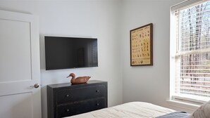A bedroom with a bed, a dresser, a flat-screen TV, a framed poster, and a window with blinds.