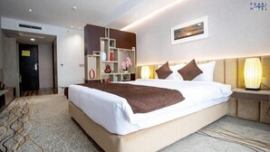 A modern hotel room with a large bed, a bedside table with a lamp, a built-in shelf unit, and a view of the cityscape. - Pearl Hotel (Dhaka)