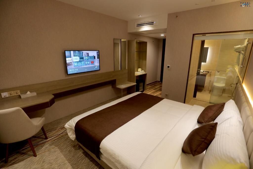 A hotel room with a bed, a desk, a TV, and a bathroom visible through a glass door.