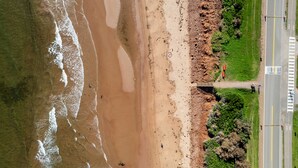 An aerial view of a beach with a road running parallel to the shore, a pedestrian path, and a small structure near the water. - Drift Wood, Spacious 2 BRs, Living, Kitchen, Full Bathroom & a Patio  (York)