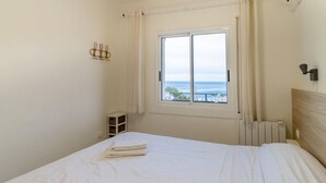 A bedroom with a bed, a window with a view of the sea, a radiator, and a small white cabinet. - Les Tonyines 67 - Apartment with views and terrace (Llança)