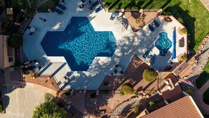 Vista aérea de un área de piscina con una piscina grande, tumbonas y un pequeño jacuzzi.