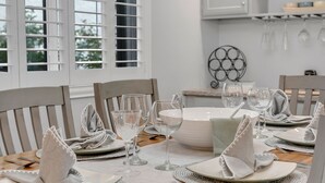 Una mesa de comedor dispuesta con platos blancos, servilletas y copas de vino, con una ventana y una cocina de fondo.