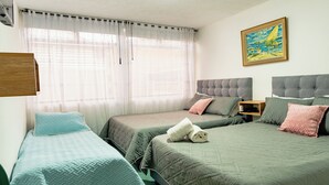 Une chambre avec deux lits simples, un grand lit, un tableau au mur et une fenêtre avec des rideaux légers.