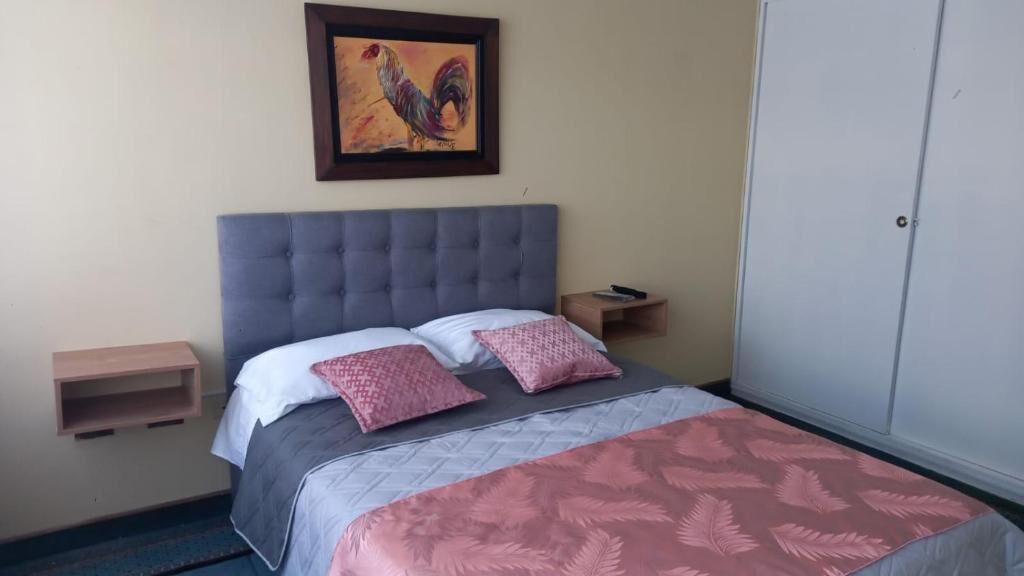 A bedroom with a tufted headboard, a bed with a patterned bedspread, two pillows, a nightstand with a lamp, and a framed artwork on the wall.