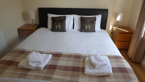 A neatly made bed with a checkered blanket and two towels, two decorative pillows with a deer design, and two bedside lamps. - Courtyard Studio (Auchendennan) (Alexandria)