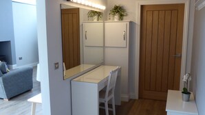 A compact, modern room with a desk, chair, and a small table with a potted plant. A wooden door and a mirror with lights are visible. - Oak Lodge (Auchendennan) - sleeps 4 guests  in 2 bedrooms (Alexandria)