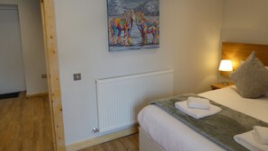 A hotel room with a bed, a painting of colorful horses, a nightstand with a lamp, and a wooden door. - The Stables Lower Cottage 5 (Auchendennan) - sleeps 2 guests  in 1 bedroom (Alexandria)