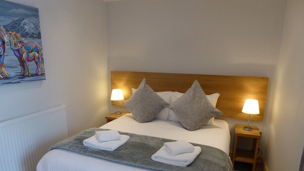 A neatly made bed with pillows and folded towels, a painting on the wall, and two bedside lamps. - The Stables Lower Cottage 5 (Auchendennan) - sleeps 2 guests  in 1 bedroom (Alexandria)