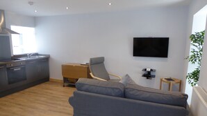 A modern living room with a grey sofa, a wooden floor, a kitchen with dark cabinets, a small wooden side table, and a flat-screen TV mounted on the wall. - The Stables Lower Cottage 5 (Auchendennan) - sleeps 2 guests  in 1 bedroom (Alexandria)