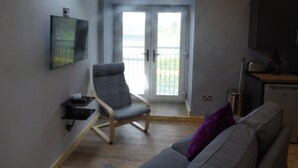 A modern living room with a grey sofa, a wooden chair, a small table, and a kitchen area with a fridge and cabinets. - The Stables Upper Cottage 7 (Auchendennan) - sleeps 2 guests  in 0 bedrooms (Alexandria)