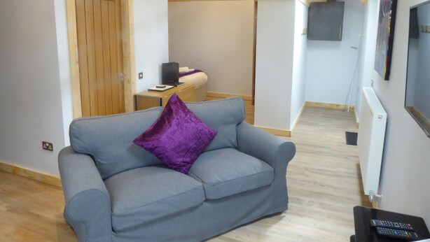 A modern living room with a grey sofa, a wooden door, a small table with a laptop, and a TV mounted on the wall. - The Stables Upper Cottage 7 (Auchendennan) - sleeps 2 guests  in 0 bedrooms (Alexandria)