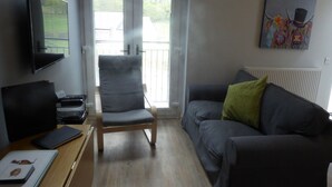 Ein Wohnzimmer mit grauem Sofa, einem grünen Kissen, einem Holztisch mit Laptop und Tablet, einem grauen Sessel und einem Bild an der Wand.
