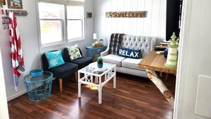 A cozy living room with a grey sofa, a small coffee table, and a blue chair. There is a sign that reads "Sand Dune" on the wall. - Sand Dune Cottage, 2 BRs, Kitchen, living and a patio (York)