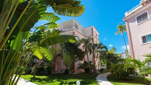 A courtyard with a paved walkway, surrounded by lush greenery and palm trees, leading to a building with a pink facade and white trim. - Cozy 1 BR apartment with AC and WiFi, walking distance to the beach in Bavaro (Punta Cana)