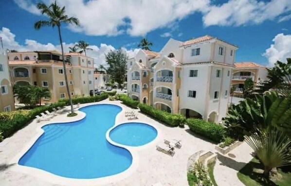 A resort with a large swimming pool, multiple buildings with balconies, and palm trees. - Cozy 1 BR apartment with AC and WiFi, walking distance to the beach in Bavaro (Punta Cana)