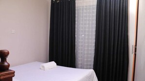A neatly made bed with a white bedspread and a rolled white towel, black curtains, and a wooden headboard. - Beautiful 1-Bed Apartment in Nairobi (Nairobi)