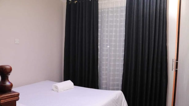 A neatly made bed with a white bedspread and a rolled white towel, black curtains, and a wooden headboard. - Beautiful 1-Bed Apartment in Nairobi (Nairobi)
