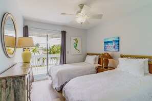 Quarto de hotel com duas camas, um ventilador de teto, uma varanda com vista para palmeiras e obras de arte nas paredes.