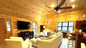 A cozy log cabin interior with a living room, TV, fireplace, and a kitchen area.
