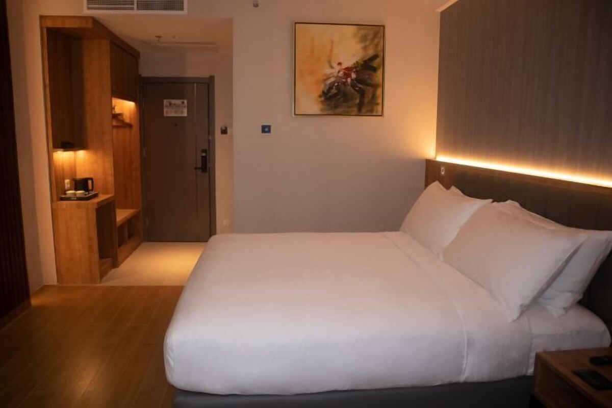 A hotel room with a large bed, a wooden wardrobe, a painting on the wall, and a nightstand with a lamp.