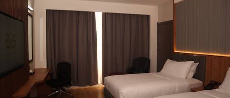 A hotel room with two beds, a desk, a chair, and a large window with curtains.