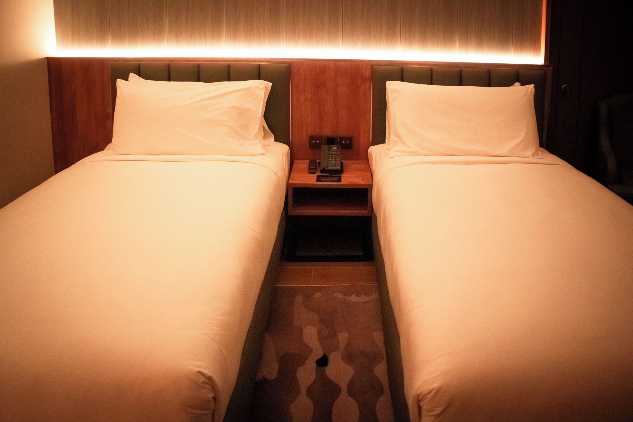 A hotel room with two single beds, a bedside table with a phone, and a headboard with built-in lighting.