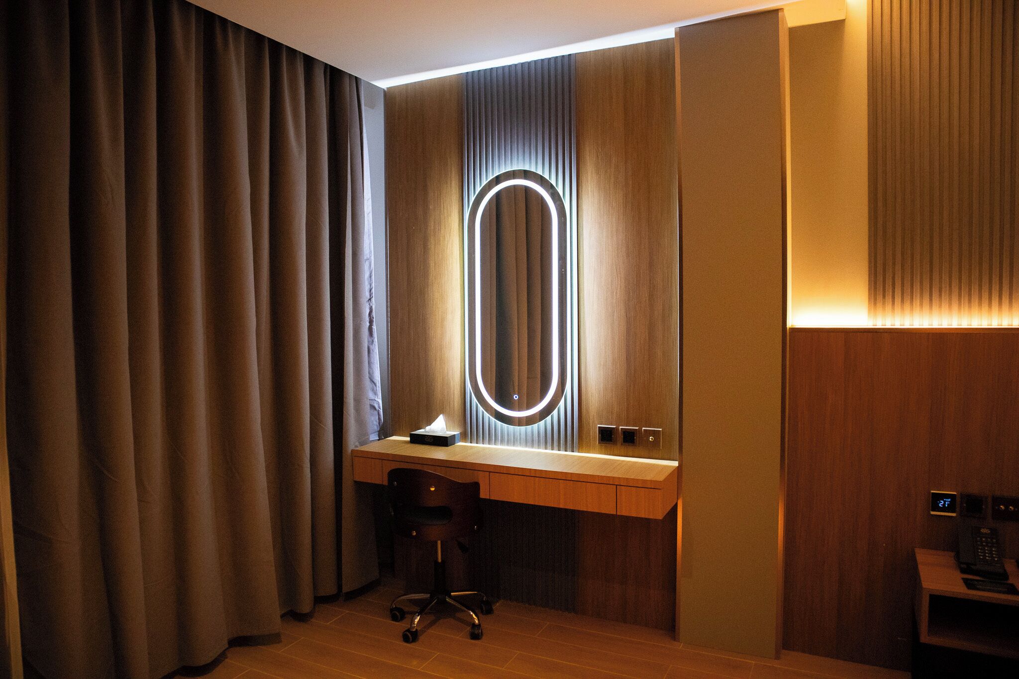 Une chambre d’hôtel avec un bureau en bois, une chaise, un grand miroir dont les bords sont éclairés, et un téléphone.