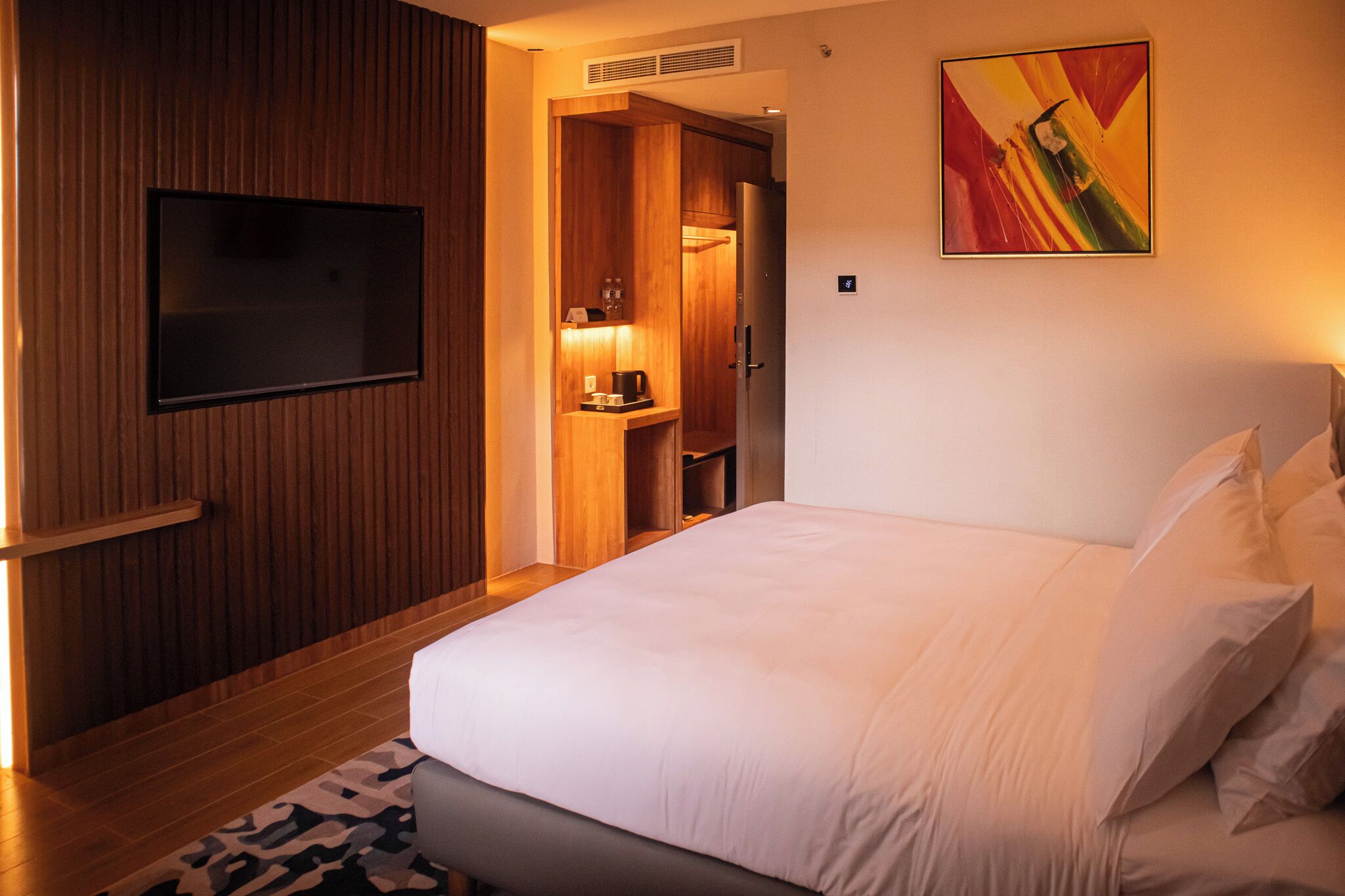 A hotel room with a bed, a television, a wooden wardrobe, and a painting on the wall.