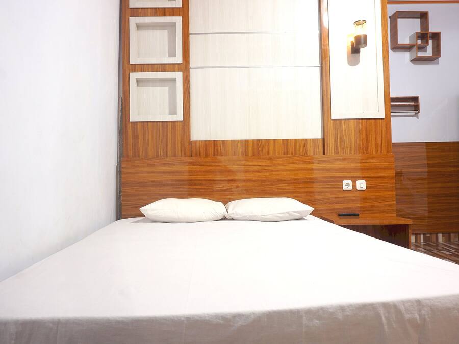 A neatly made bed with white linens and two pillows, a wooden headboard with built-in shelves, and a bedside table with a remote control.