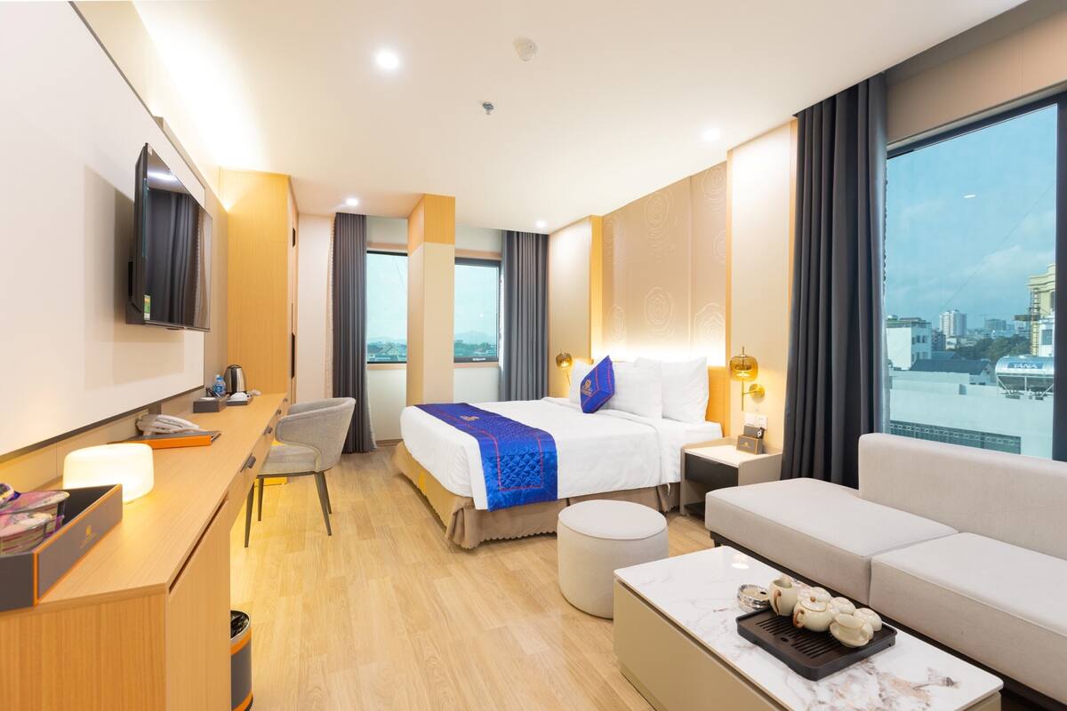 Ein Hotelzimmer mit Bett, Schreibtisch, Sofa, Fernseher und einem Fenster mit Blick auf die Stadt.