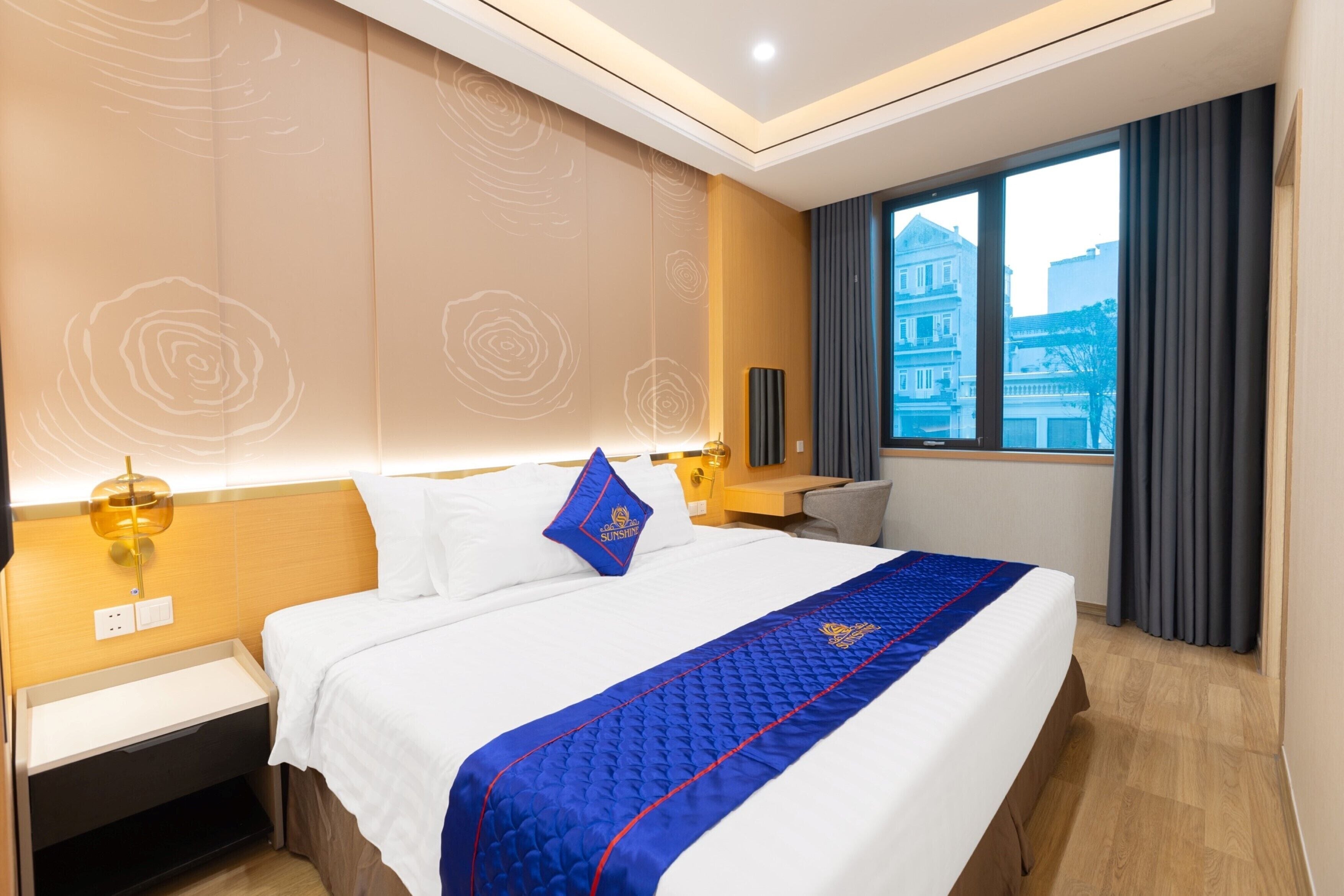 A hotel room with a large bed, a nightstand, a lamp, and a view of a building through a window.