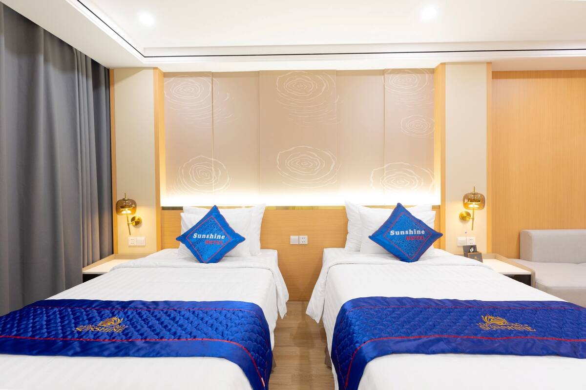 Ein Hotelzimmer mit zwei Betten, jeweils mit blauer Bettdecke und einem Kopfkissen mit dem Namen des Hotels. Zwei Nachttische mit Lampen, ein hölzernes Kopfteil und ein großes Fenster mit Blick.
