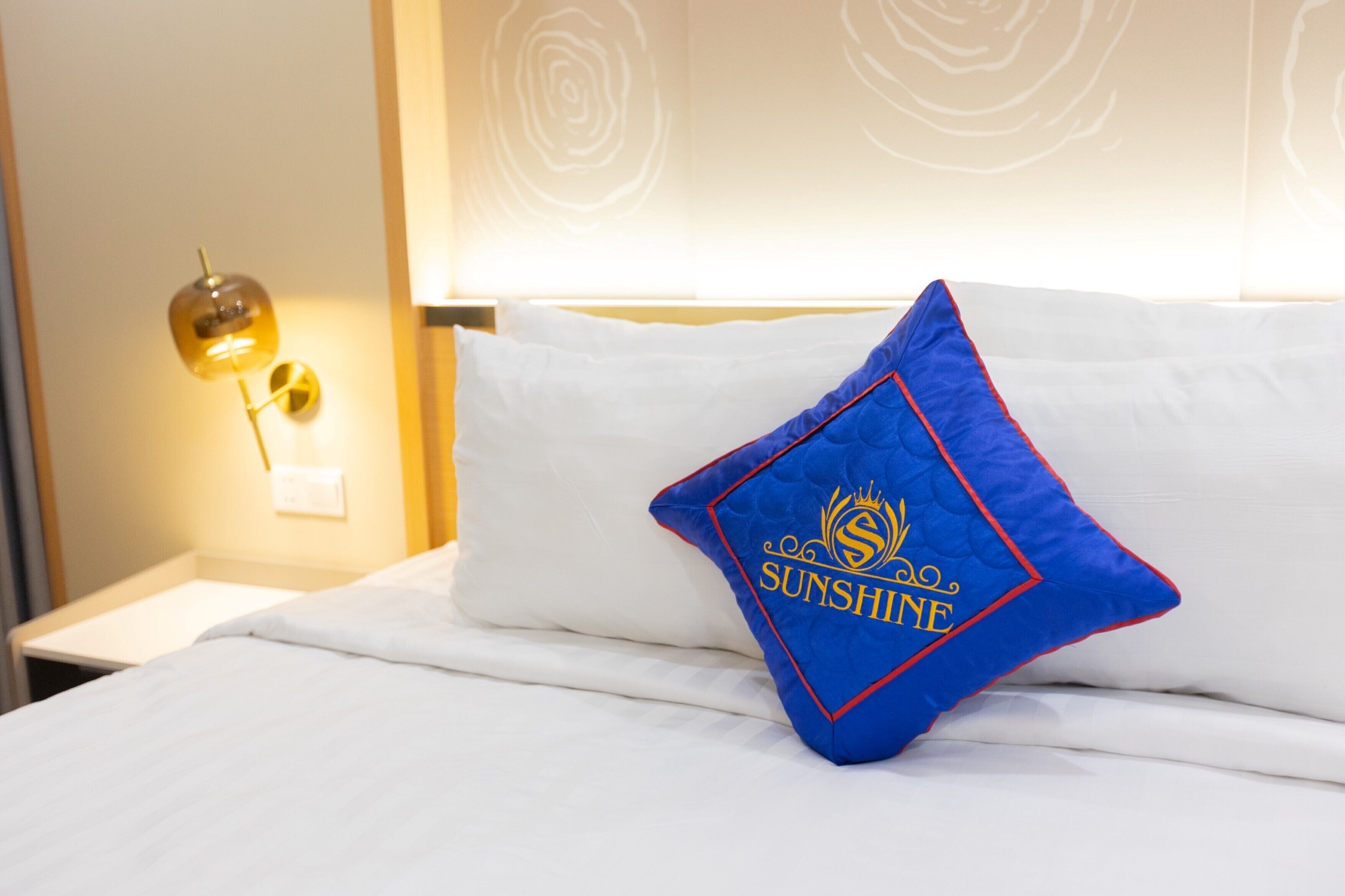 A neatly made bed with a white comforter and a blue pillow with "SUNSHINE" embroidered on it.