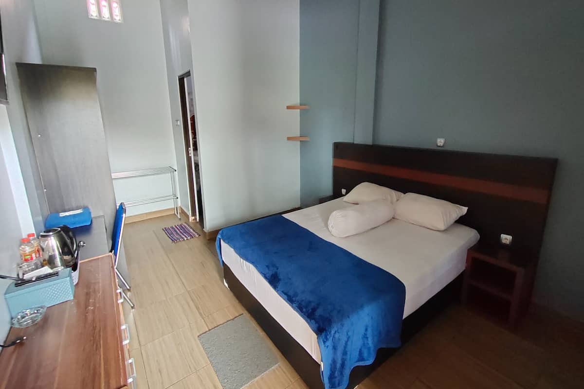 Una habitación de hotel con una cama, una mesita con una tetera y una caja azul, una silla y suelo de madera.