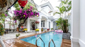 A swimming pool with wooden deck, surrounded by potted plants and flowers, and a white building with balconies. - Phuc Villa (Da Nang)
