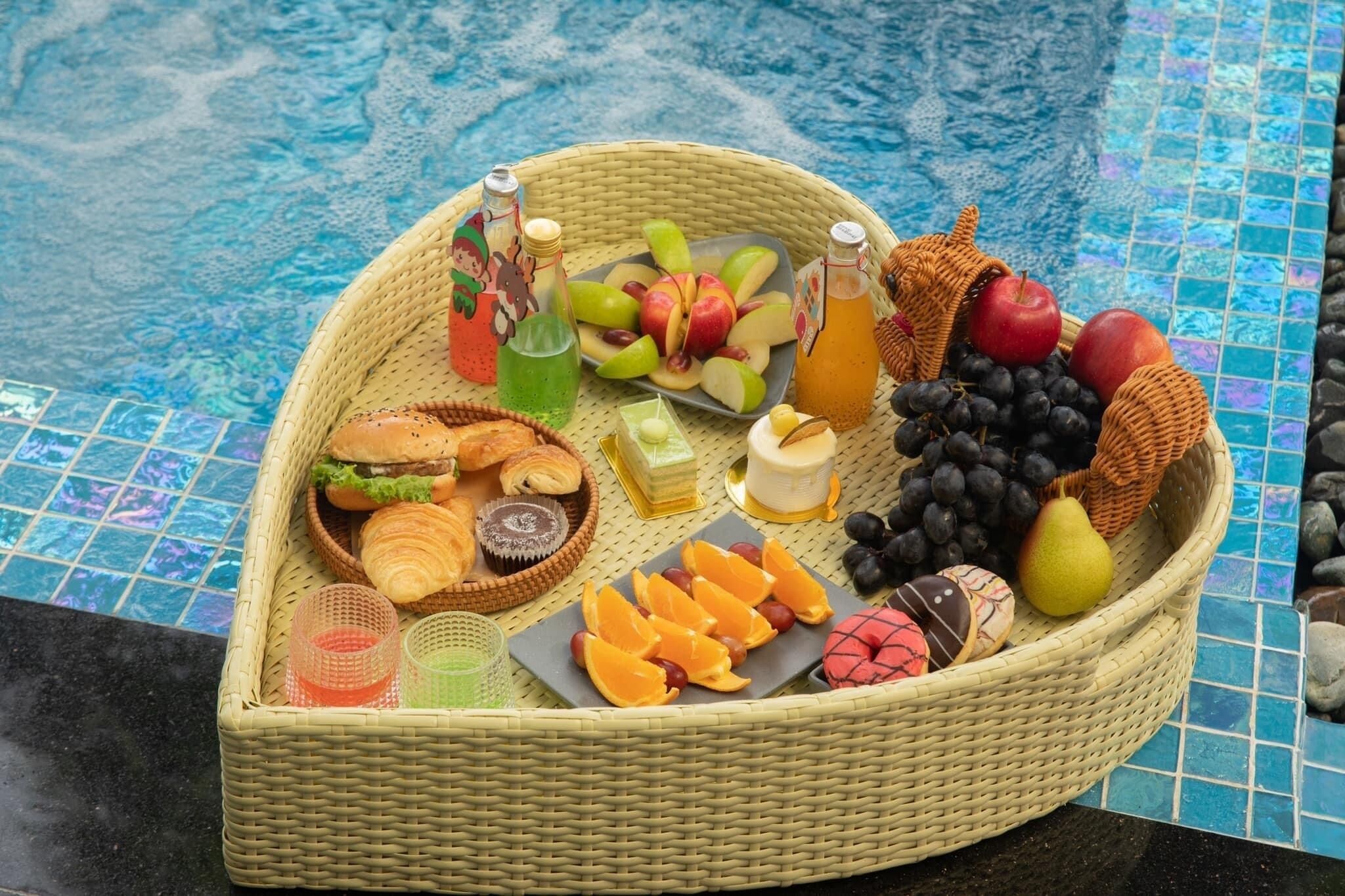 A wicker basket filled with various fruits, pastries, and beverages by a poolside.