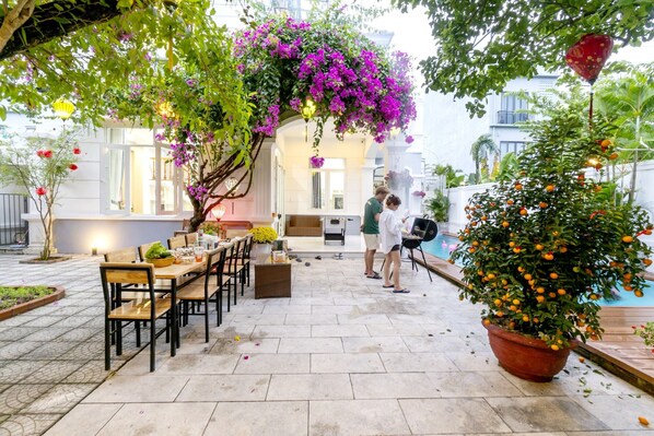 Un área de comedor al aire libre con piscina, rodeada de exuberante follaje y plantas florecientes.